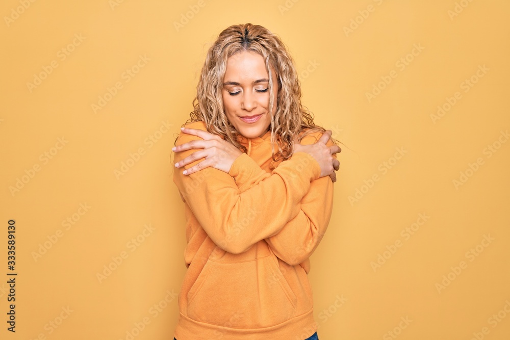 Young beautiful blonde sporty woman wearing casual sweatshirt over yellow background Hugging oneself happy and positive, smiling confident. Self love and self care Young beautiful blonde sporty woman wearing casual sweatshirt over yellow background Hugging oneself happy and positive, smiling confident. Self love and self care