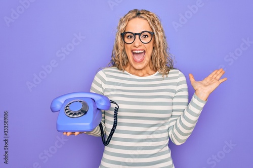 Young beautiful blonde woman holding vintage telephone over isolated purple background celebrating victory with happy smile and winner expression with raised hands