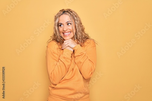 Young beautiful blonde sporty woman wearing casual sweatshirt over yellow background laughing nervous and excited with hands on chin looking to the side