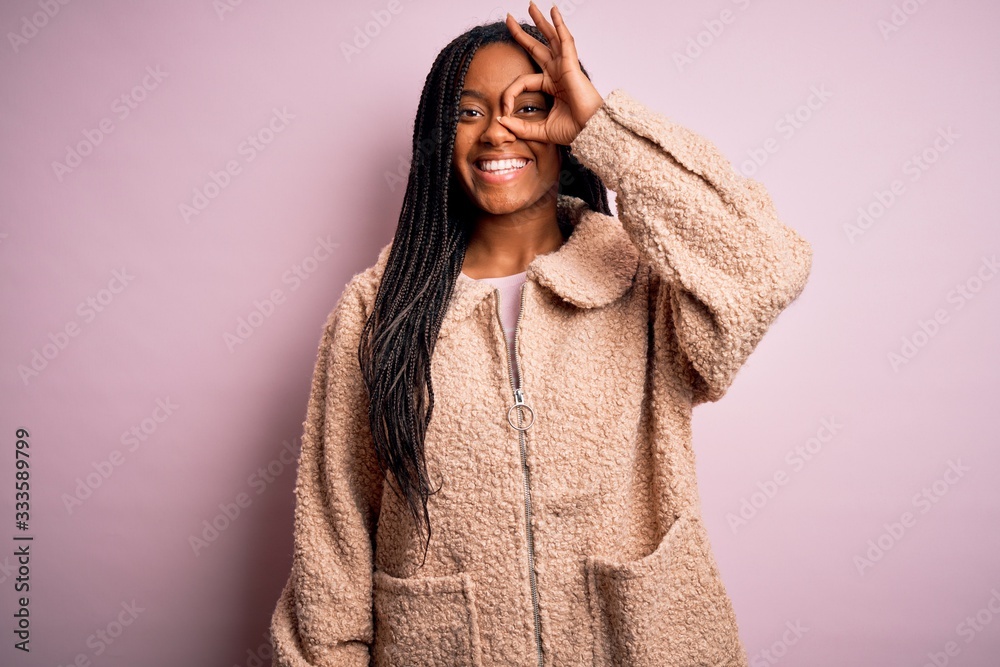 Young african american woman wearing fashion winter coat over pink isolated background doing ok gesture with hand smiling, eye looking through fingers with happy face. Young african american woman wearing fashion winter coat over pink isolated background doing ok gesture with hand smiling, eye looking through fingers with happy face.