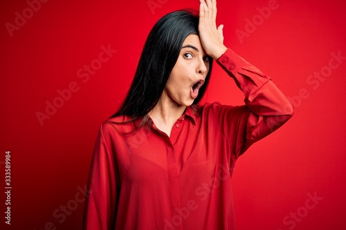 Young beautiful brunette woman wearing casual shirt standing over red background surprised with hand on head for mistake, remember error. Forgot, bad memory concept.