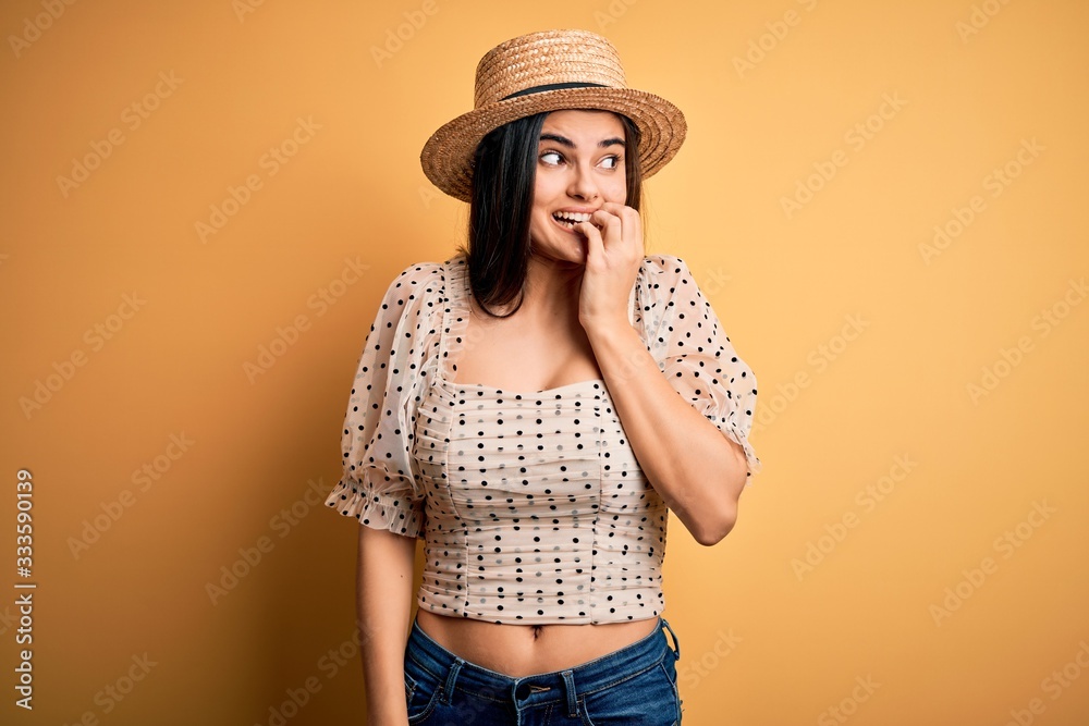 Young beautiful brunette woman wearing t-shirt and summer hat over yellow background looking stressed and nervous with hands on mouth biting nails. Anxiety problem. Young beautiful brunette woman wearing t-shirt and summer hat over yellow background looking stressed and nervous with hands on mouth biting nails. Anxiety problem.