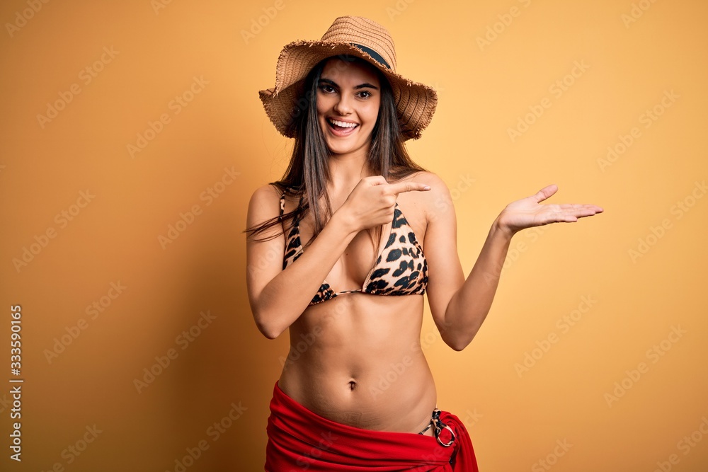 Young beautiful brunette woman on vacation wearing swimsuit and summer hat amazed and smiling to the camera while presenting with hand and pointing with finger. Young beautiful brunette woman on vacation wearing swimsuit and summer hat amazed and smiling to the camera while presenting with hand and pointing with finger.