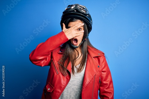 Young beautiful brunette motorcycliste woman wearing motorcycle helmet and jacket peeking in shock covering face and eyes with hand, looking through fingers with embarrassed expression. © Krakenimages.com