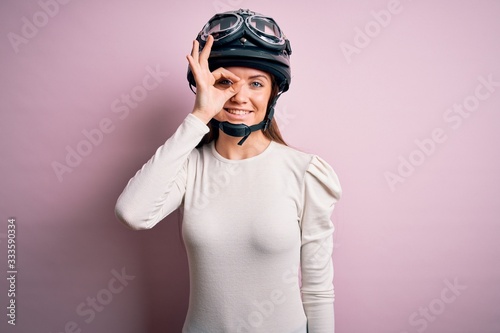 Young beautiful motorcyclist woman with blue eyes wearing moto helmet over pink background doing ok gesture with hand smiling, eye looking through fingers with happy face.