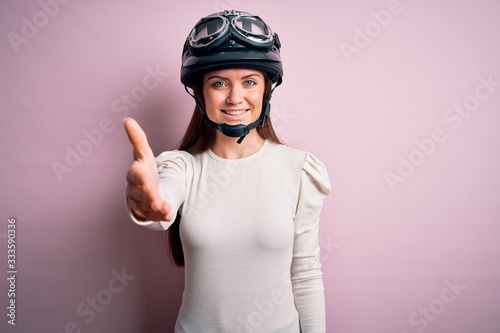 Young beautiful motorcyclist woman with blue eyes wearing moto helmet over pink background smiling friendly offering handshake as greeting and welcoming. Successful business.
