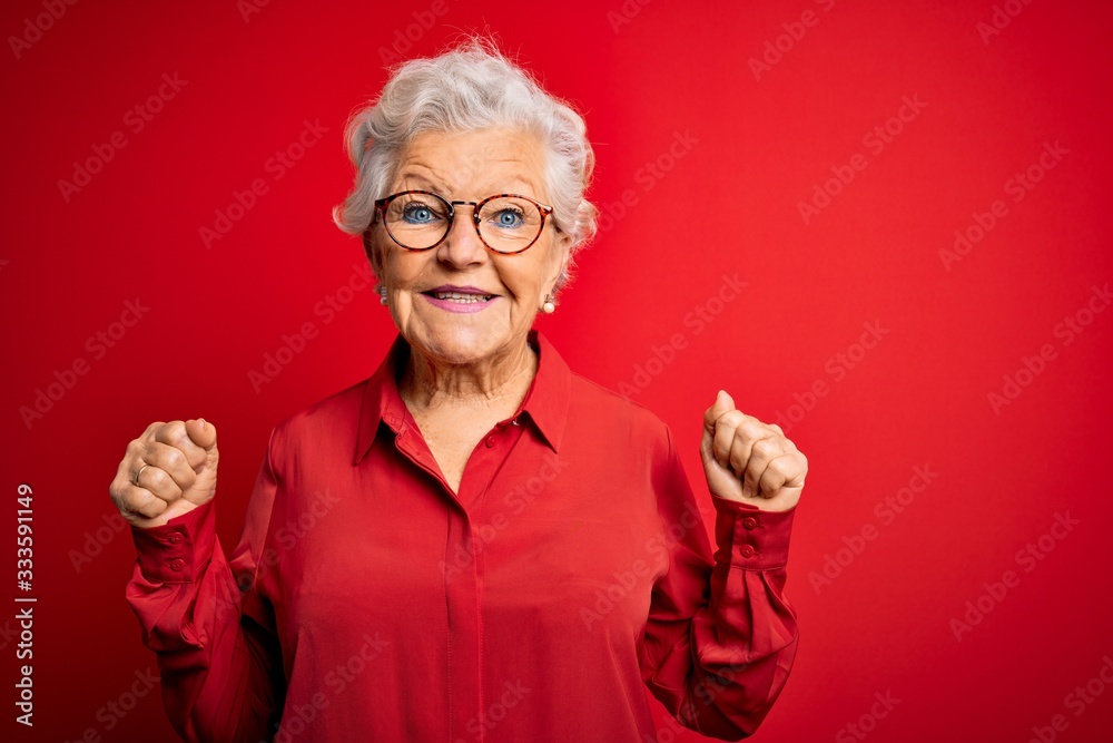 Senior beautiful grey-haired woman wearing casual shirt and glasses over red background celebrating surprised and amazed for success with arms raised and open eyes. Winner concept. Senior beautiful grey-haired woman wearing casual shirt and glasses over red background celebrating surprised and amazed for success with arms raised and open eyes. Winner concept.