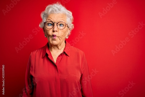Senior beautiful grey-haired woman wearing casual shirt and glasses over red background making fish face with lips, crazy and comical gesture. Funny expression. © Krakenimages.com