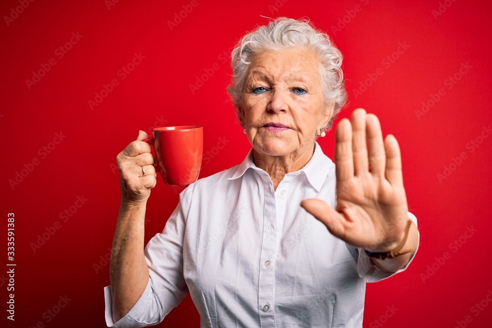 Senior beautiful woman drinking mug of coffee standing over isolated red background with open hand doing stop sign with serious and confident expression, defense gesture Senior beautiful woman drinking mug of coffee standing over isolated red background with open hand doing stop sign with serious and confident expression, defense gesture