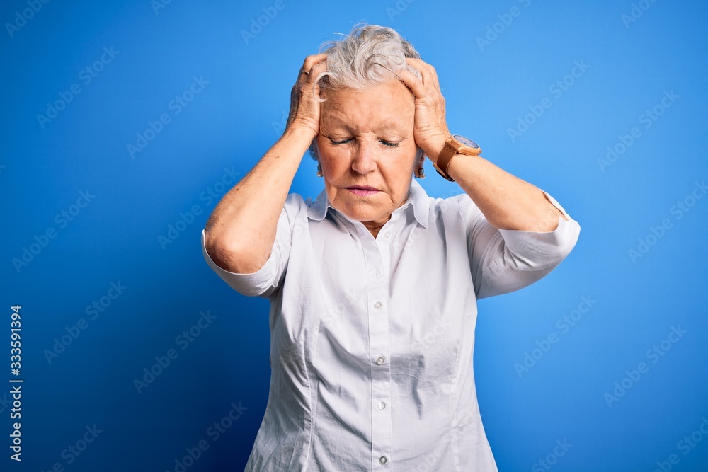 Senior beautiful woman wearing elegant shirt standing over isolated blue background suffering from headache desperate and stressed because pain and migraine. Hands on head. Senior beautiful woman wearing elegant shirt standing over isolated blue background suffering from headache desperate and stressed because pain and migraine. Hands on head.