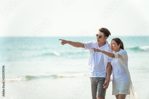 Smiling young Asian couple man and beautiful woman holding hands together and walking on the beach in summertime. Healthy family people enjoying relax lifestyle in romantic summer holiday vacation.