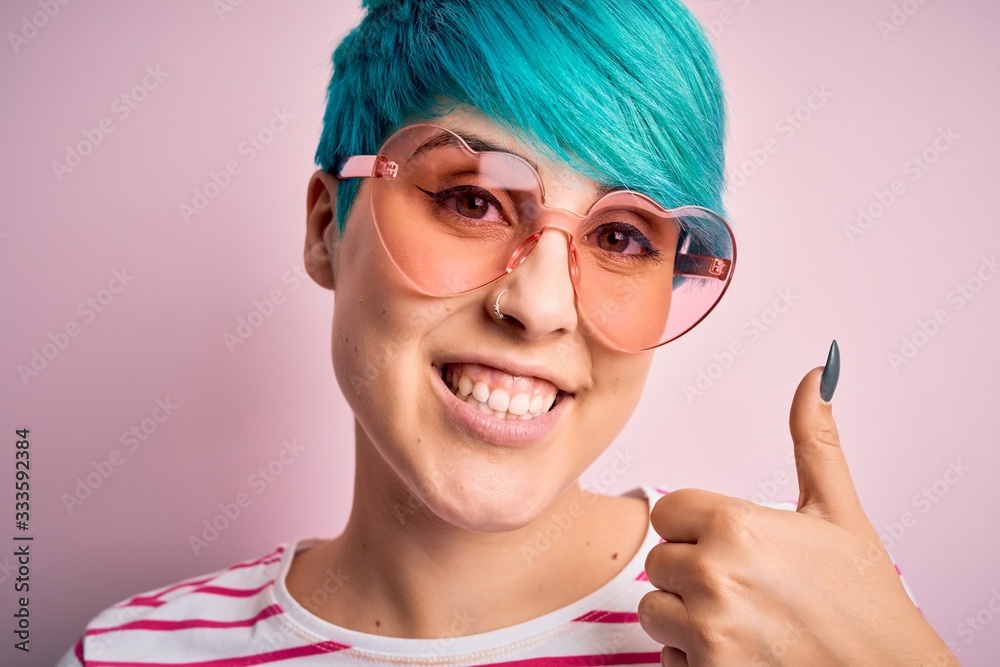 Young beautiful woman with blue fashion hair wearing fanny glasses with hearts happy with big smile doing ok sign, thumb up with fingers, excellent sign