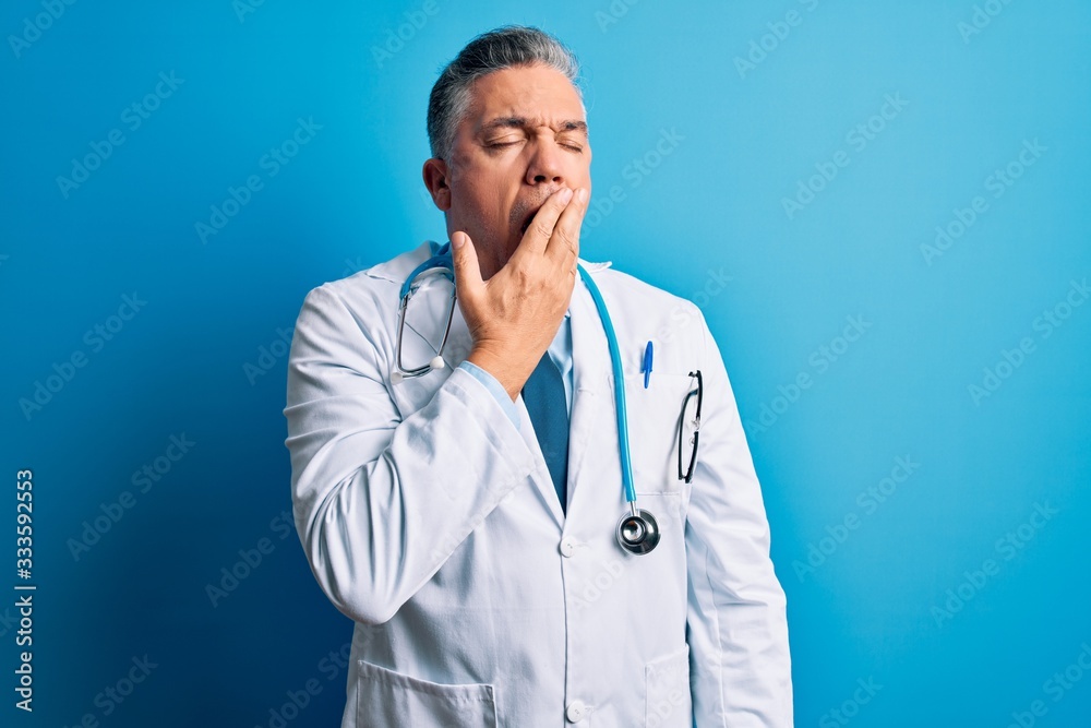 Middle age handsome grey-haired doctor man wearing coat and blue stethoscope bored yawning tired covering mouth with hand. Restless and sleepiness. Middle age handsome grey-haired doctor man wearing coat and blue stethoscope bored yawning tired covering mouth with hand. Restless and sleepiness.