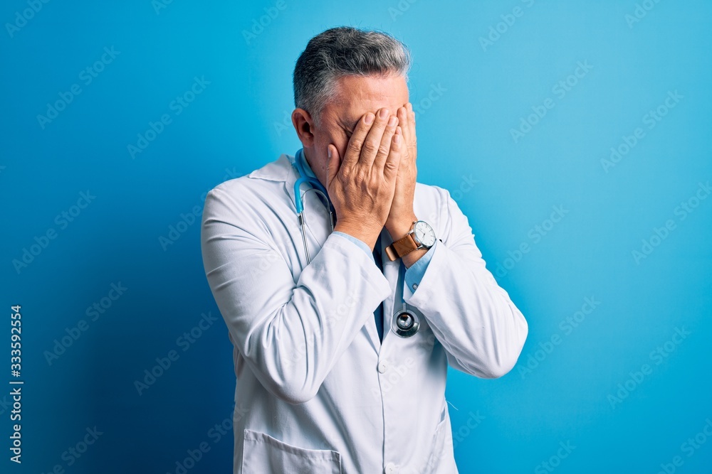 Middle age handsome grey-haired doctor man wearing coat and blue stethoscope with sad expression covering face with hands while crying. Depression concept. Middle age handsome grey-haired doctor man wearing coat and blue stethoscope with sad expression covering face with hands while crying. Depression concept.