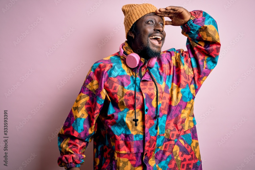 Young handsome african american man wearing colorful coat and cap over pink background very happy and smiling looking far away with hand over head. Searching concept.