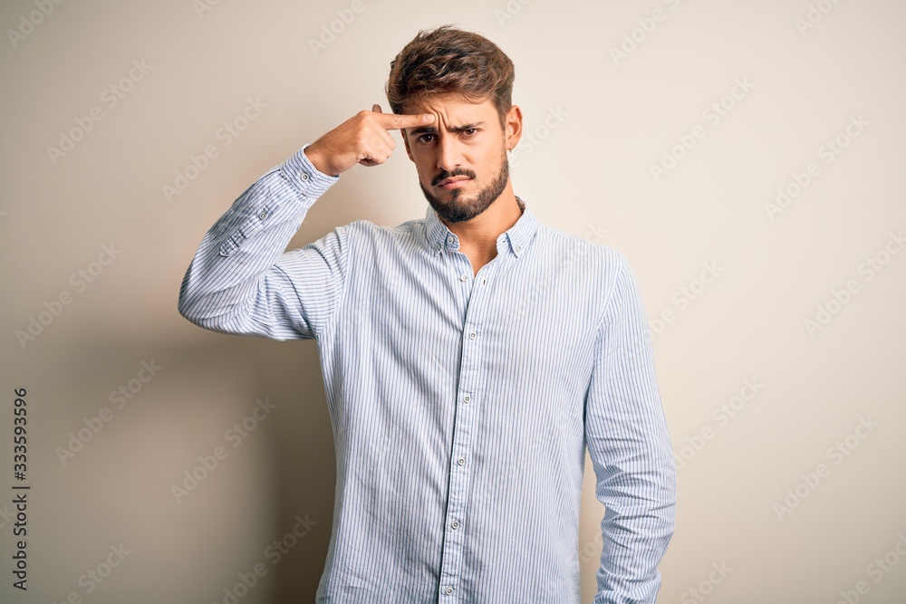 Young handsome man with beard wearing striped shirt standing over white background pointing unhappy to pimple on forehead, ugly infection of blackhead. Acne and skin problem Young handsome man with beard wearing striped shirt standing over white background pointing unhappy to pimple on forehead, ugly infection of blackhead. Acne and skin problem