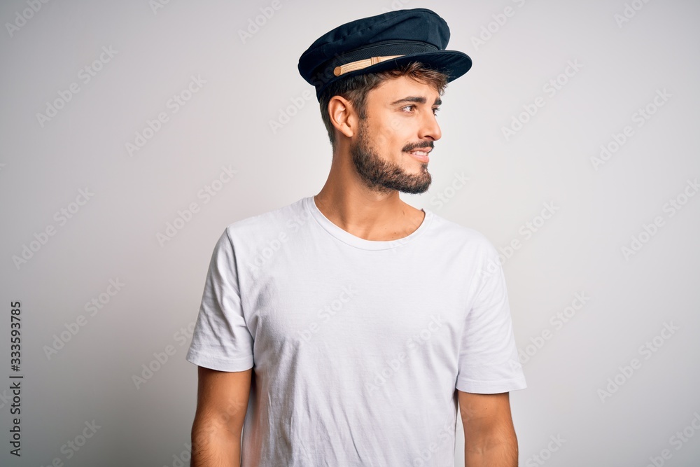 Young driver man with beard wearing hat standing over isolated white background looking away to side with smile on face, natural expression. Laughing confident. Young driver man with beard wearing hat standing over isolated white background looking away to side with smile on face, natural expression. Laughing confident.