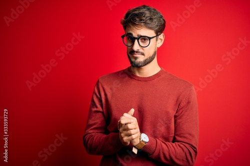 Young handsome man with beard wearing glasses and sweater standing over red background with hands together and crossed fingers smiling relaxed and cheerful. Success and optimistic
