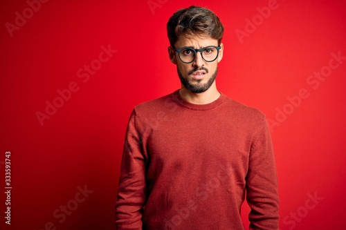 Young handsome man with beard wearing glasses and sweater standing over red background In shock face, looking skeptical and sarcastic, surprised with open mouth