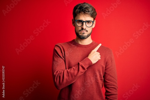 Young handsome man with beard wearing glasses and sweater standing over red background Pointing with hand finger to the side showing advertisement, serious and calm face