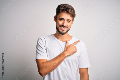 Young handsome man with beard wearing casual t-shirt standing over white background cheerful with a smile of face pointing with hand and finger up to the side with happy and natural expression on face