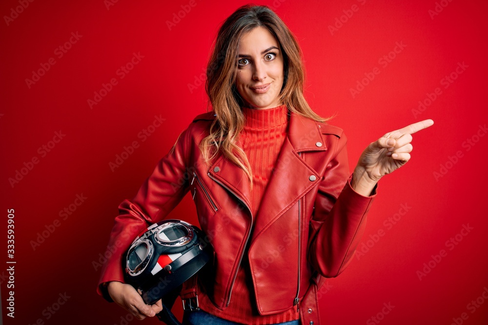 Naklejka premium Beautiful brunette motorcyclist woman wearing red jacket holding motorcycle helmet very happy pointing with hand and finger to the side