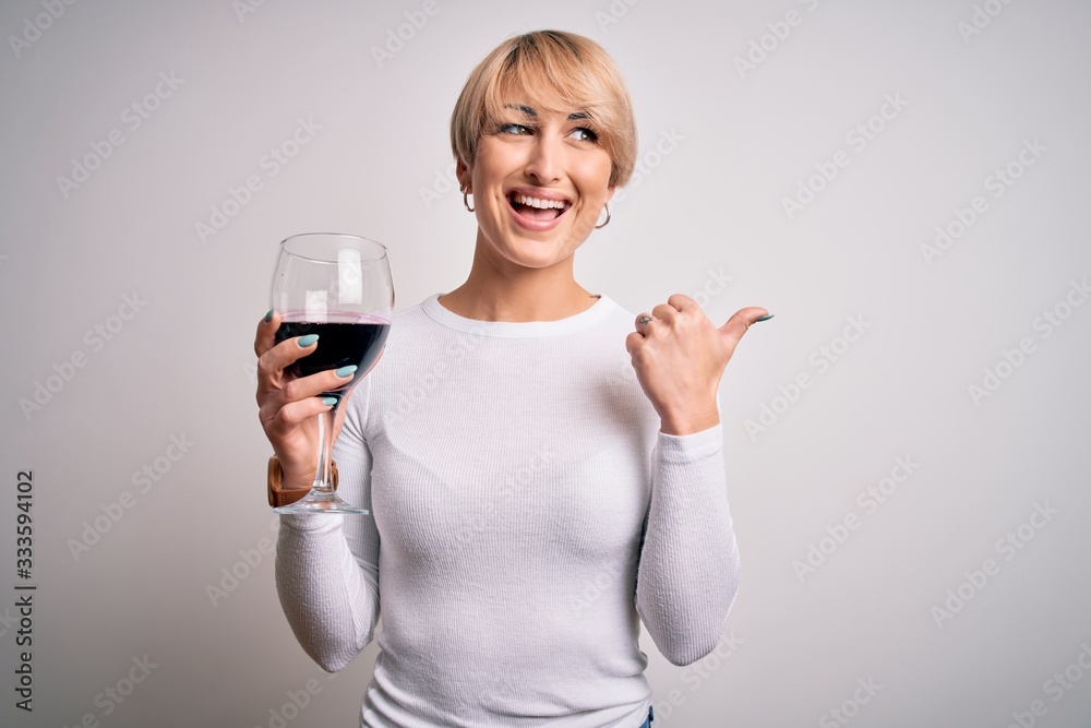 Young blonde woman with short hair drinking a glass of red wine over isolated background pointing and showing with thumb up to the side with happy face smiling Young blonde woman with short hair drinking a glass of red wine over isolated background pointing and showing with thumb up to the side with happy face smiling