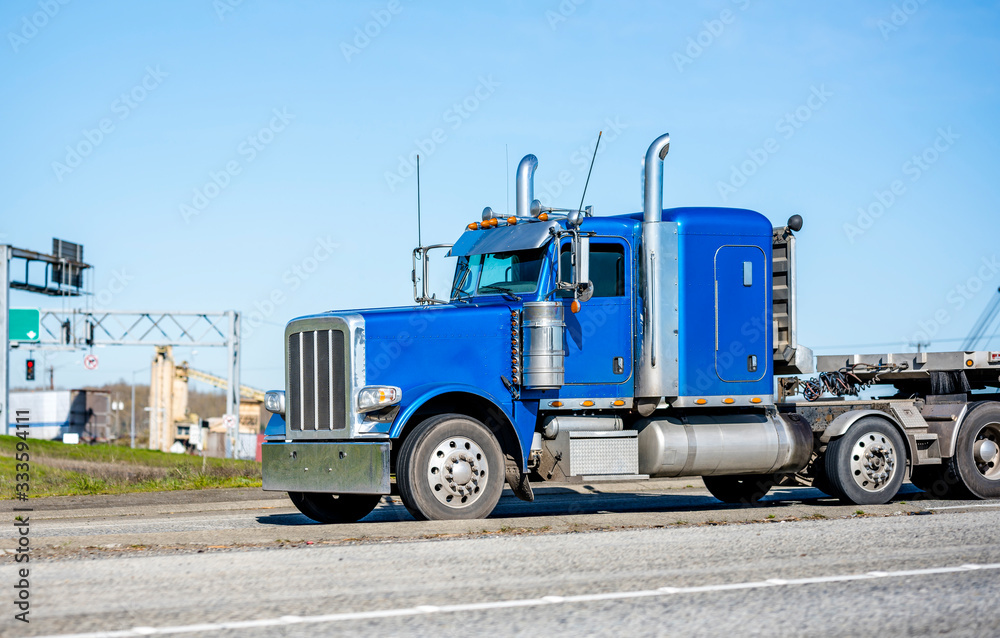 Classic blue big rig semi truck with tall exhaust pipes transporting cargo on flat bed semi trailer running on the road in industrial area
