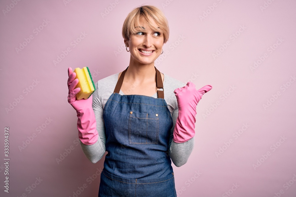 Young blonde cleaner woman with short hair wearing apron and gloves holding scrub scourer pointing and showing with thumb up to the side with happy face smiling Young blonde cleaner woman with short hair wearing apron and gloves holding scrub scourer pointing and showing with thumb up to the side with happy face smiling
