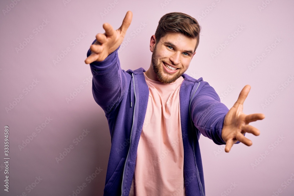 Naklejka premium Young blond man with beard and blue eyes wearing purple sweatshirt over pink background looking at the camera smiling with open arms for hug. Cheerful expression embracing happiness.