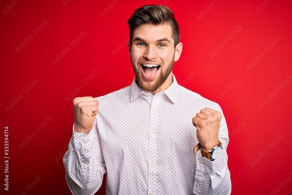 Young blond businessman with beard and blue eyes wearing elegant shirt over red background celebrating surprised and amazed for success with arms raised and open eyes. Winner concept. Young blond businessman with beard and blue eyes wearing elegant shirt over red background celebrating surprised and amazed for success with arms raised and open eyes. Winner concept.