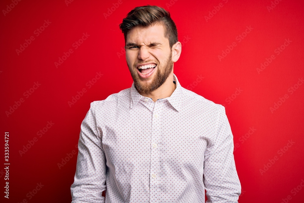 Young blond businessman with beard and blue eyes wearing elegant shirt over red background winking looking at the camera with sexy expression, cheerful and happy face. Young blond businessman with beard and blue eyes wearing elegant shirt over red background winking looking at the camera with sexy expression, cheerful and happy face.
