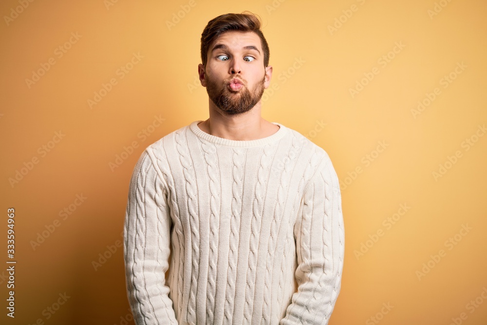 Young blond man with beard and blue eyes wearing white sweater over yellow background making fish face with lips, crazy and comical gesture. Funny expression. Young blond man with beard and blue eyes wearing white sweater over yellow background making fish face with lips, crazy and comical gesture. Funny expression.