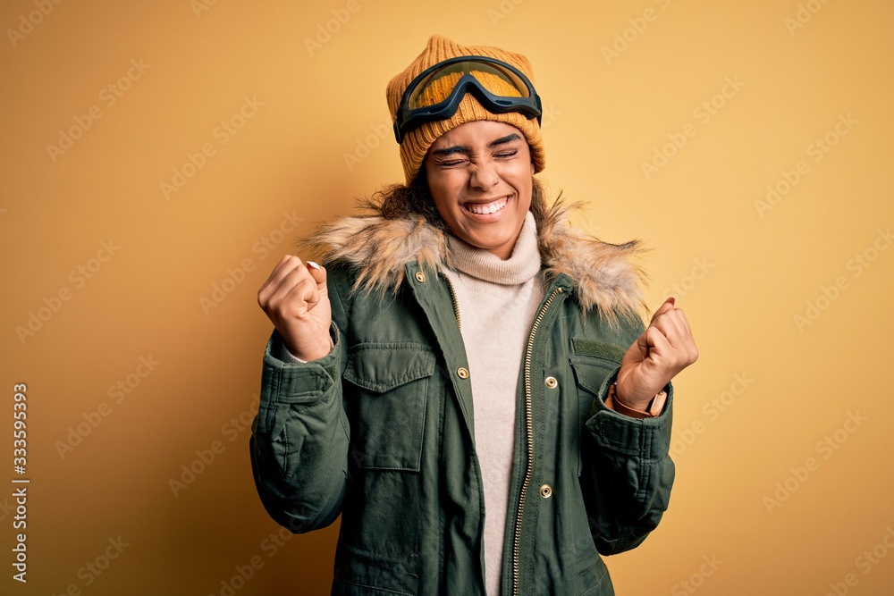 Fototapeta premium Young african american afro skier girl wearing snow sportswear and ski goggles very happy and excited doing winner gesture with arms raised, smiling and screaming for success. Celebration concept.
