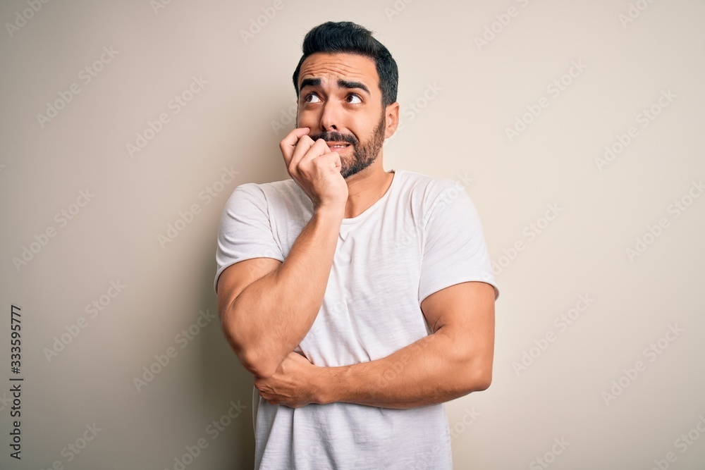 Young handsome man with beard wearing casual t-shirt standing over white background looking stressed and nervous with hands on mouth biting nails. Anxiety problem. Young handsome man with beard wearing casual t-shirt standing over white background looking stressed and nervous with hands on mouth biting nails. Anxiety problem.