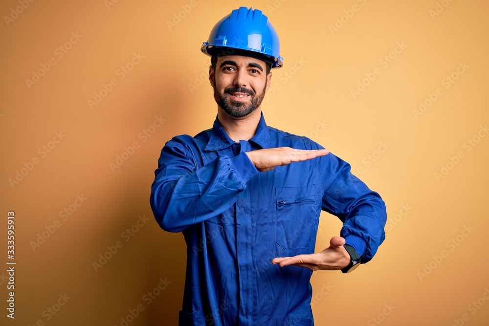 Mechanic man with beard wearing blue uniform and safety helmet over yellow background gesturing with hands showing big and large size sign, measure symbol. Smiling looking at the camera. Measuring 