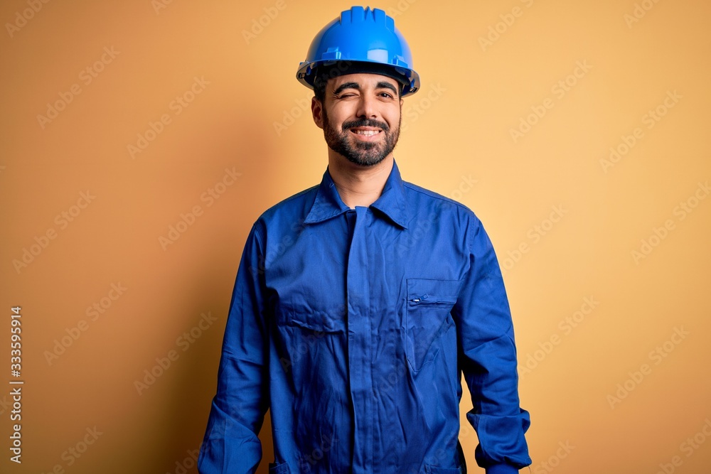 Mechanic man with beard wearing blue uniform and safety helmet over yellow background winking looking at the camera with sexy expression, cheerful and happy face.