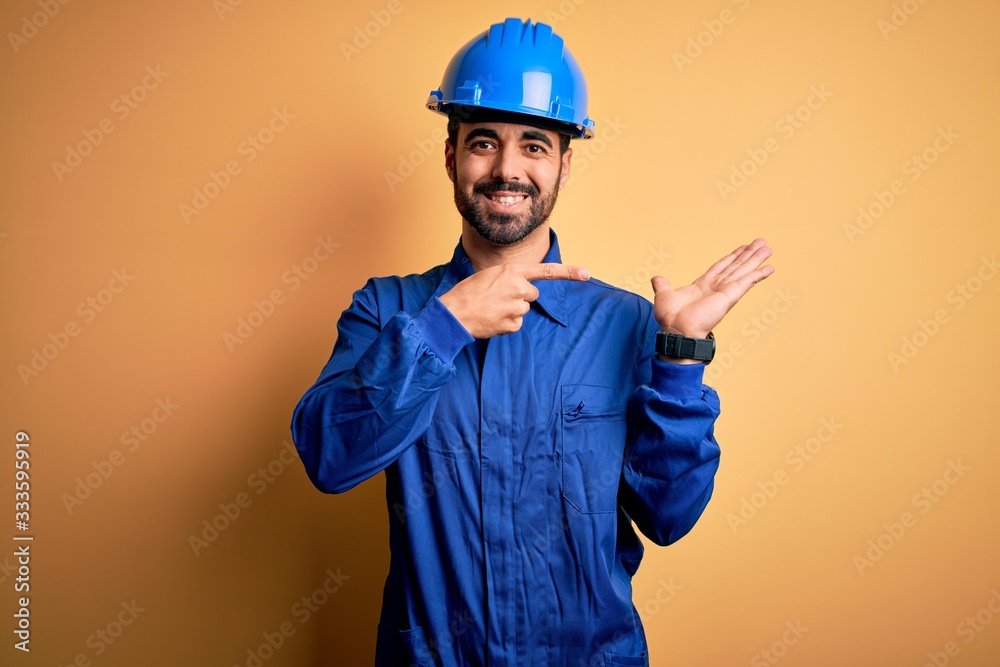 Mechanic man with beard wearing blue uniform and safety helmet over yellow background amazed and smiling to the camera while presenting with hand and pointing with finger.