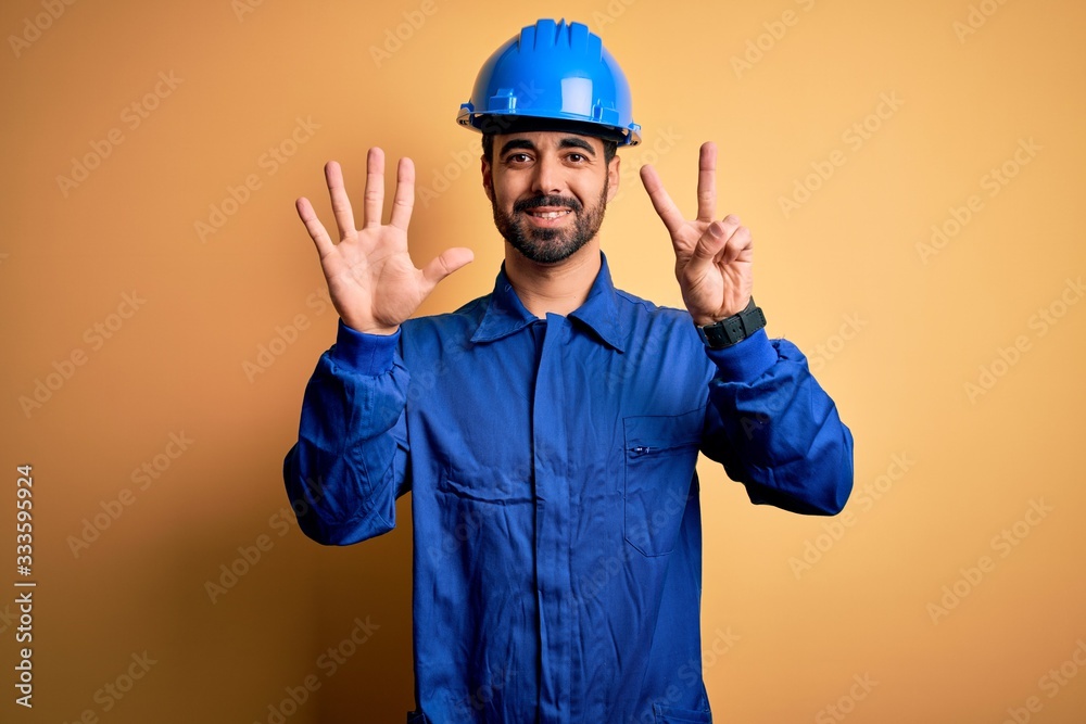 Mechanic man with beard wearing blue uniform and safety helmet over yellow background showing and pointing up with fingers number seven while smiling confident and happy.