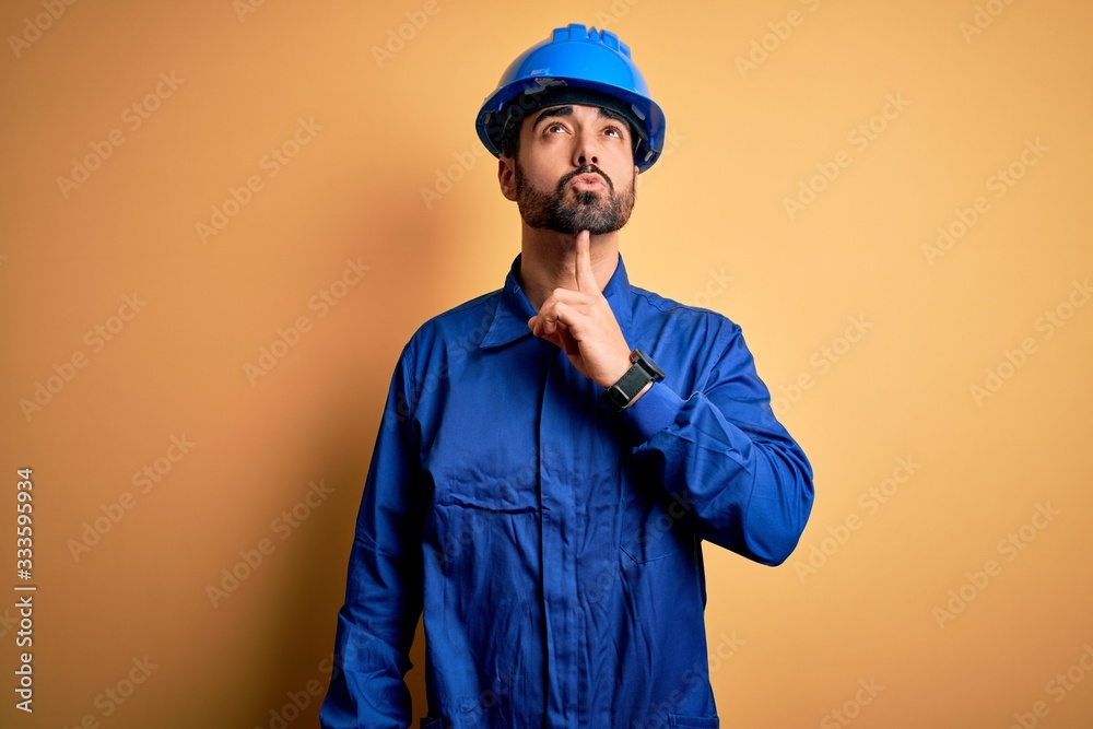 Mechanic man with beard wearing blue uniform and safety helmet over yellow background Thinking concentrated about doubt with finger on chin and looking up wondering