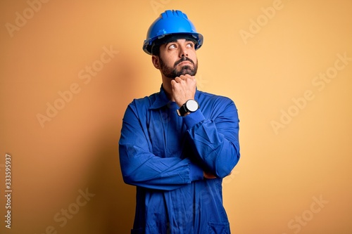 Mechanic man with beard wearing blue uniform and safety helmet over yellow background with hand on chin thinking about question, pensive expression. Smiling with thoughtful face. Doubt concept.
