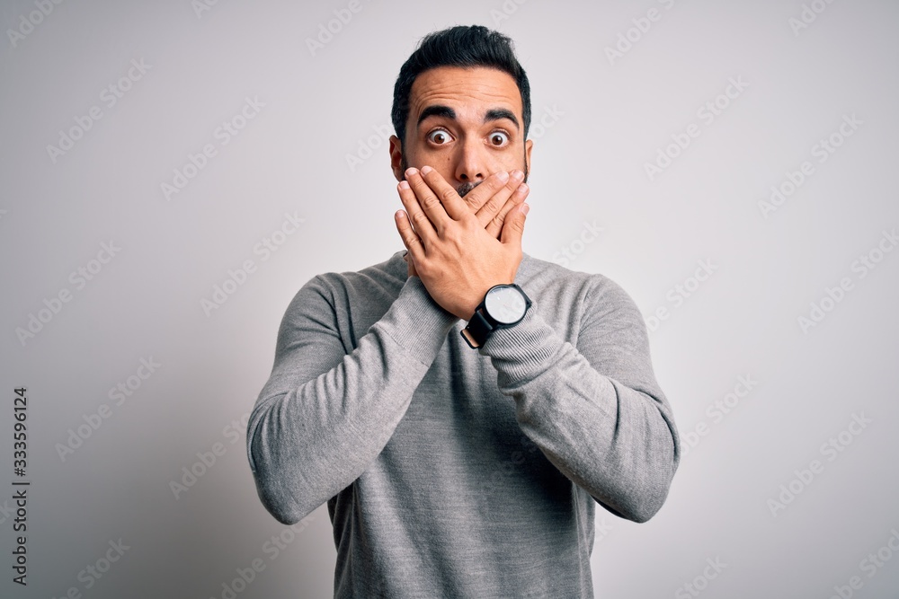 Young handsome man with beard wearing casual sweater standing over white background shocked covering mouth with hands for mistake. Secret concept.