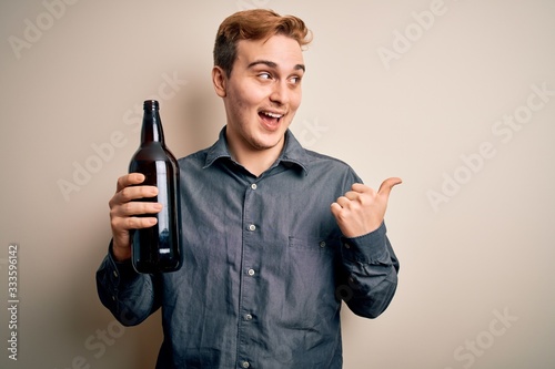 Young handsome redhead man drinking bottle of beer over isolated white background pointing thumb up to the side smiling happy with open mouth