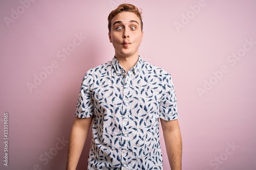 Young handsome redhead man wearing casual summer shirt standing over pink background making fish face with lips, crazy and comical gesture. Funny expression.
