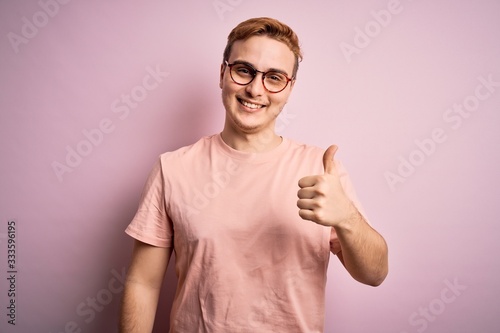 Young handsome redhead man wearing casual t-shirt standing over isolated pink background doing happy thumbs up gesture with hand. Approving expression looking at the camera showing success. © Krakenimages.com