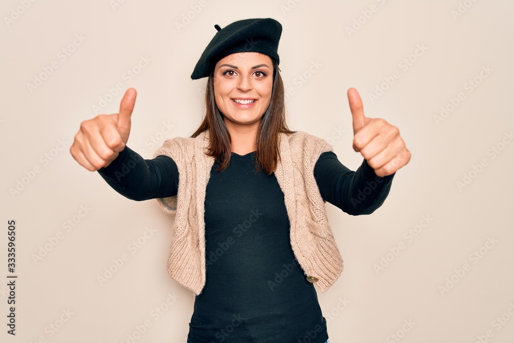 Young beautiful brunette woman wearing casual french beret over white background approving doing positive gesture with hand, thumbs up smiling and happy for success. Winner gesture. Young beautiful brunette woman wearing casual french beret over white background approving doing positive gesture with hand, thumbs up smiling and happy for success. Winner gesture.