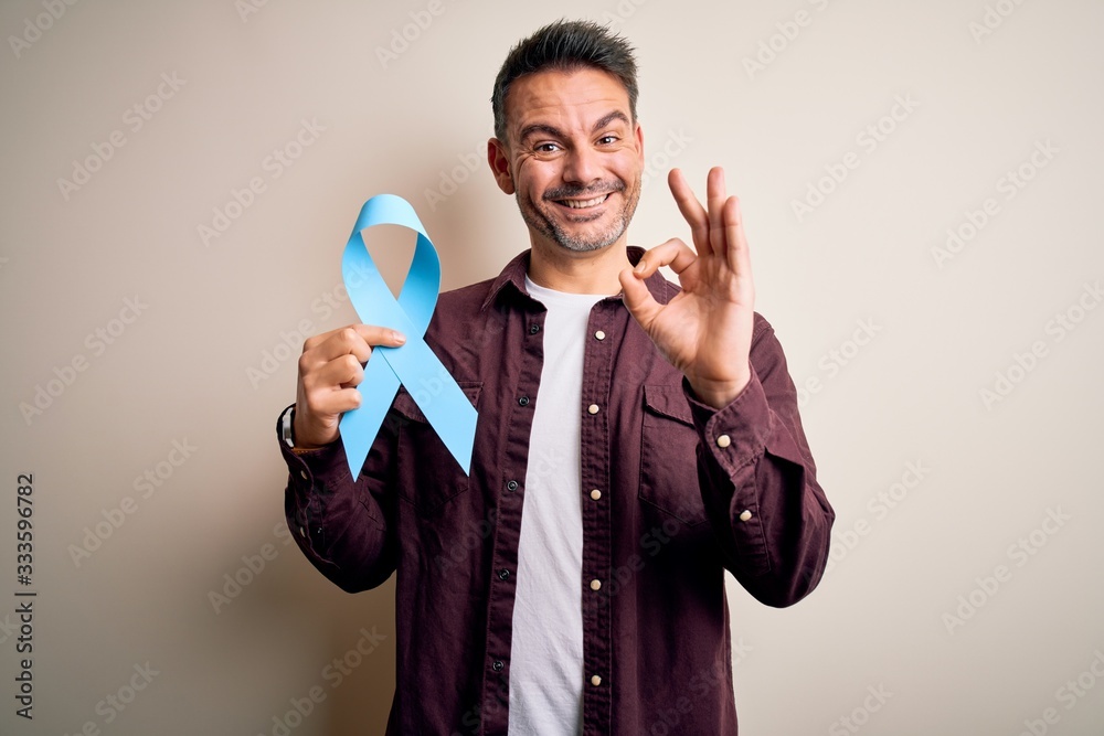 Young handsome man holding blue cancer ribbon over isolated white background doing ok sign with fingers, excellent symbol Young handsome man holding blue cancer ribbon over isolated white background doing ok sign with fingers, excellent symbol