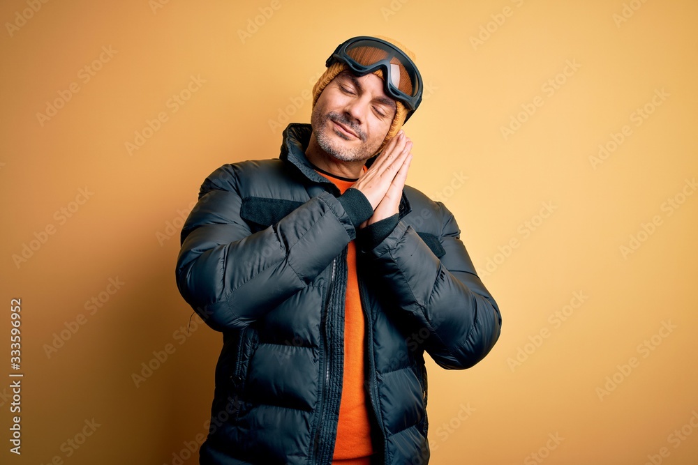 Young handsome skier man skiing wearing snow sportswear using ski goggles sleeping tired dreaming and posing with hands together while smiling with closed eyes. Young handsome skier man skiing wearing snow sportswear using ski goggles sleeping tired dreaming and posing with hands together while smiling with closed eyes.