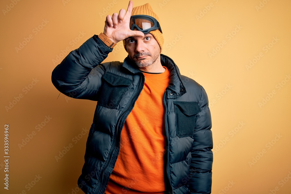 Young handsome skier man skiing wearing snow sportswear using ski goggles making fun of people with fingers on forehead doing loser gesture mocking and insulting.