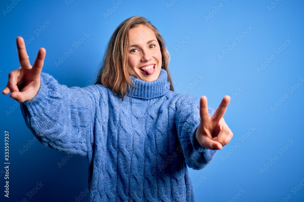 Young beautiful blonde woman wearing casual turtleneck sweater over blue background smiling with tongue out showing fingers of both hands doing victory sign. Number two.